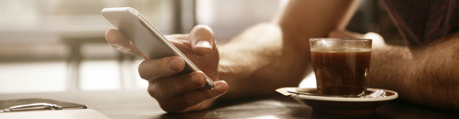 Hand mit Mobilfon, im Vordergrund eine Tasse Kaffee